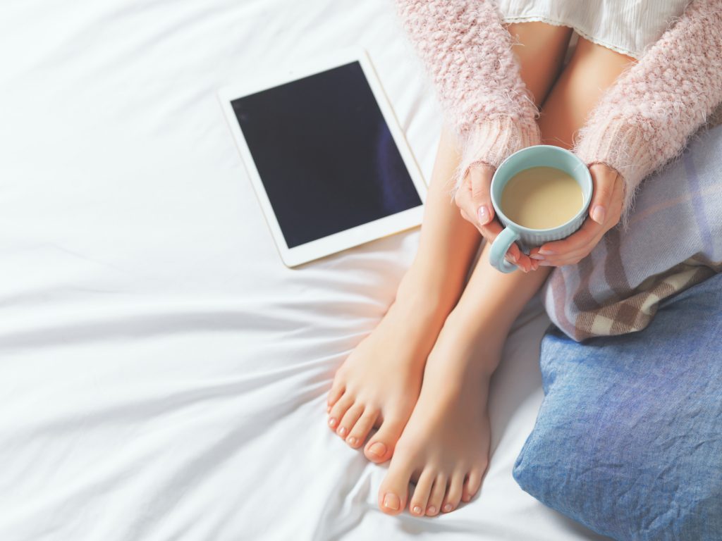 Woman using tablet at cozy home atmosphere on the bed. Young beautiful woman enjoying free time using technological device, holding a cup of cocoa or coffee. Soft light